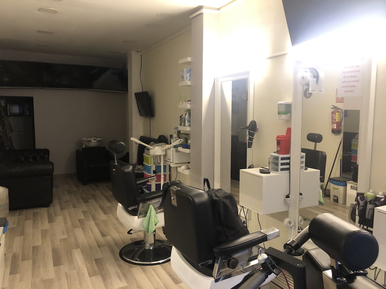 Interior vacío de una peluquería: suelo de madera clara, tres sillones de barbero negros, estanterías con productos, un sofá negro al fondo y un lavacabezas junto a la pared.